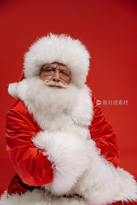 据说脾气暴躁的圣诞老人穿着舞台服装，双臂交叉站在红色背景上