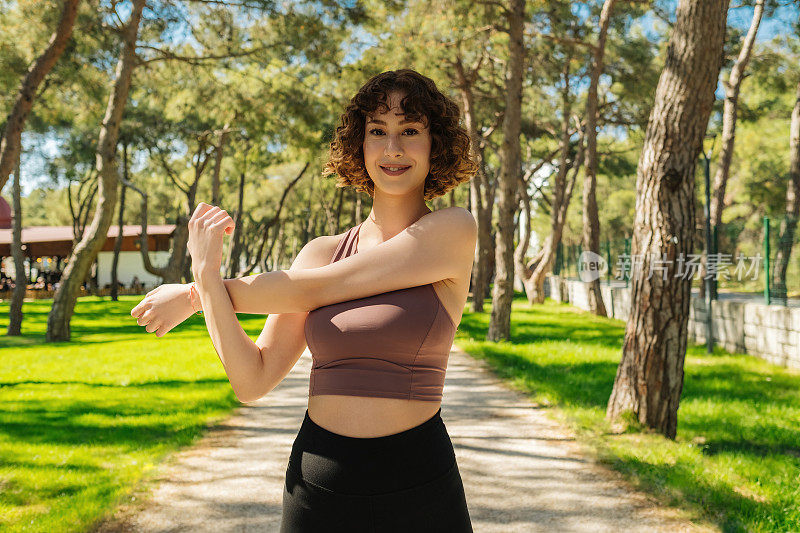 一个微笑的健康的红发女人伸展手臂在城市公园，户外的肖像。健身女士锻炼，热身，伸展手臂。