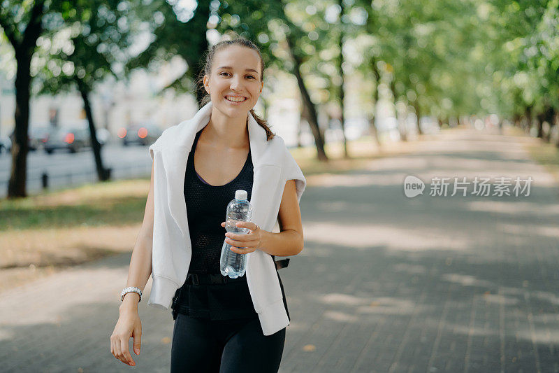 户外拍摄的高兴运动的女人喝新鲜的水从瓶子在夏天走绿色的城市公园，脸上有牙齿的微笑，引导健康的生活方式。训练后恢复水的平衡