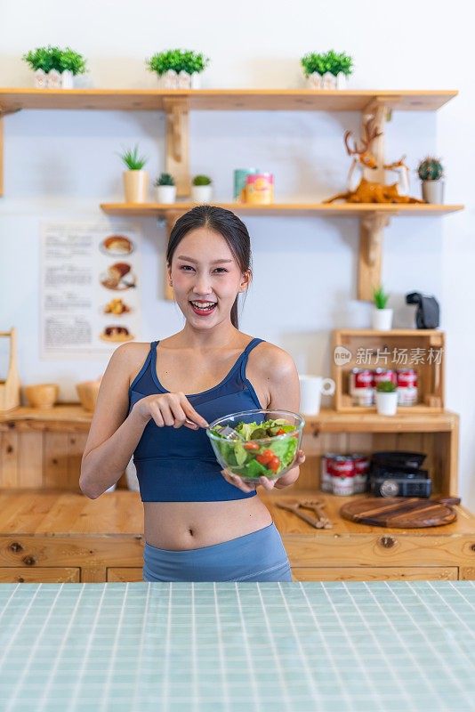 年轻美丽的亚洲女性女士的照片在运动服快乐地享受吃沙拉和樱桃番茄在她的房子的厨房节食和减肥健康的生活方式