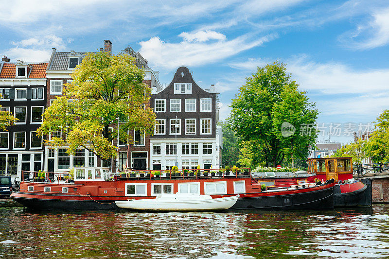 在一个阳光灿烂的日子里，停泊在阿姆斯特丹中心运河边的船屋。蓝天白云为背景。