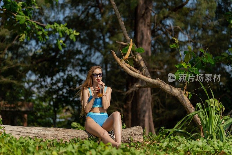 美丽的白人女性戴着太阳镜和蓝色比基尼，在海滩旁郁郁葱葱的热带树木公园里喝着混合冰饮料