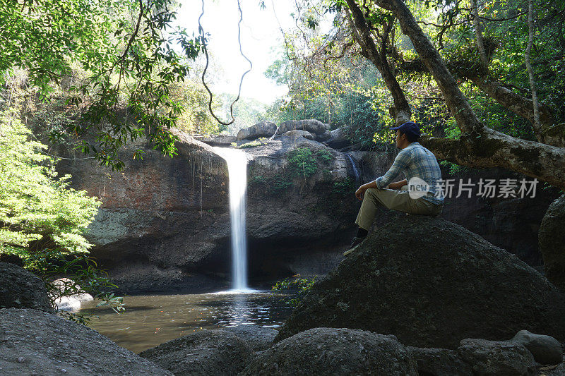 亚洲男人坐在有瀑布背景的岩石上。