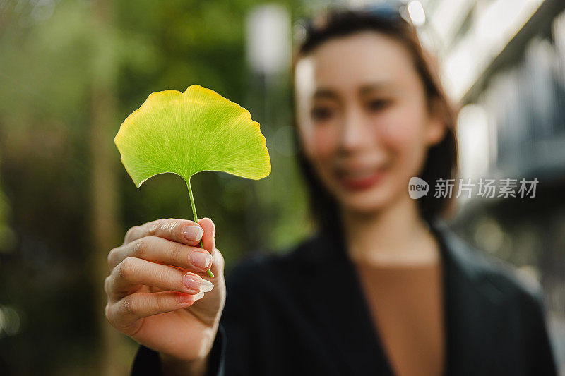 一个迷人的女人站在城市的街道上，手里拿着一片银杏叶。