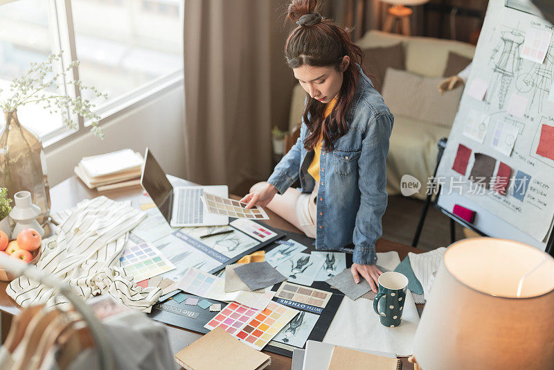 智能手机上的新工作咨询亚洲成年女性时装设计师布裁缝工作的新系列素描，选择新的色块面料样品在她的办公桌在她的设计工作室家庭工作室
