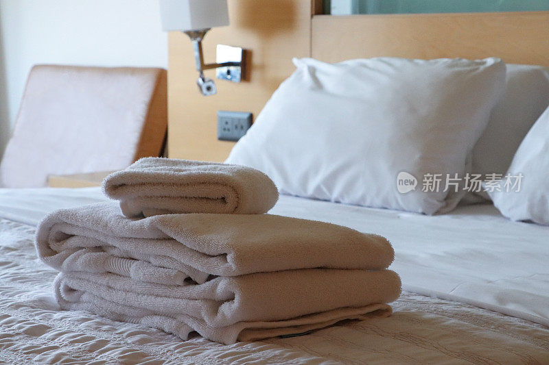 特写图像的堆叠白色酒店毛巾坐在白色羽绒被床罩，折叠浴床单和手巾，枕头靠在木床头板，床头柜，壁灯，皮椅，重点在前景