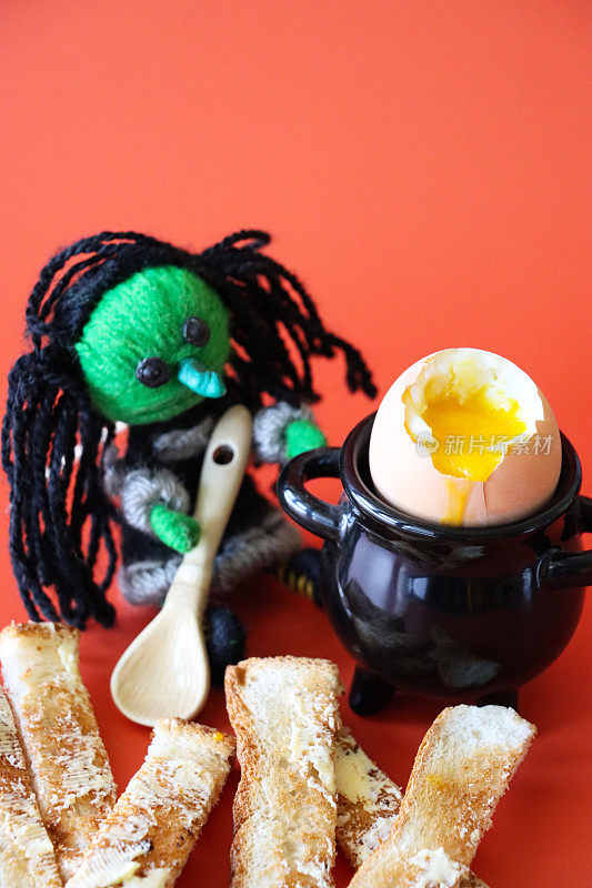 万圣节主题早餐图片黑色大锅蛋杯包含软煮蛋与士兵和绿色羊毛女巫与木长勺，溏心蛋黄，白面包吐司片与黄油，橙色背景，高视图