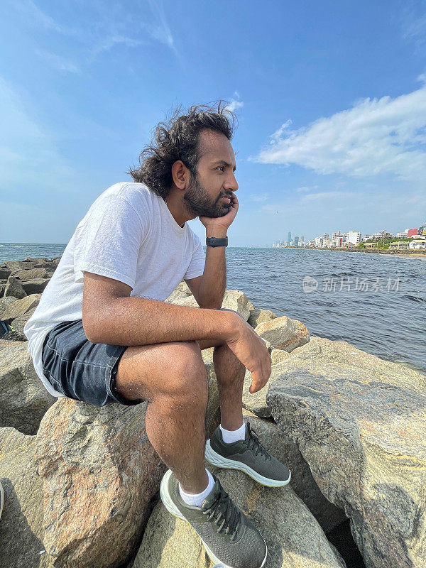 特写图片，印度男子坐在手里拿着下巴，在斯里兰卡海岸线上的人造岩石海防上看着风景，海岸侵蚀抛石装甲墙，海岸管理，大海，背景是斯里兰卡科伦坡的城市摩天大楼