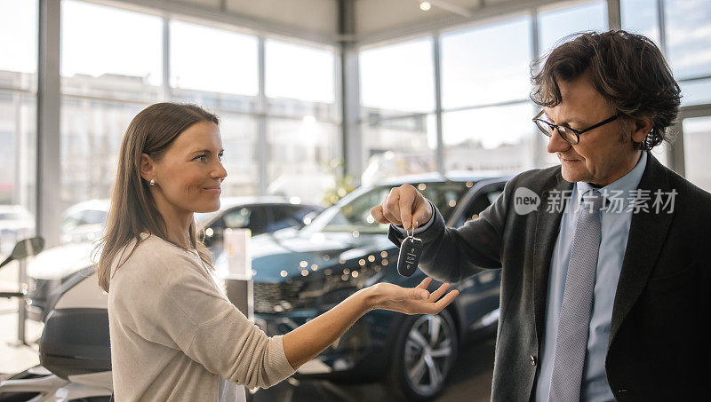 图为，一名女子在汽车经销店的室内接受新车钥匙时，露出满意的表情看着一名男汽车销售员