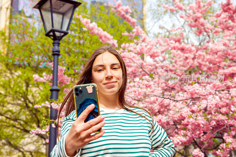 女子打视频电话，用手机自拍。年轻微笑的欧洲白人女性在樱花树的背景下，户外公园。
