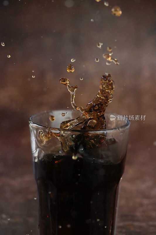 特写图片:冰块被扔进汽水杯，碳酸饮料飞溅，棕色背景，焦点在前景