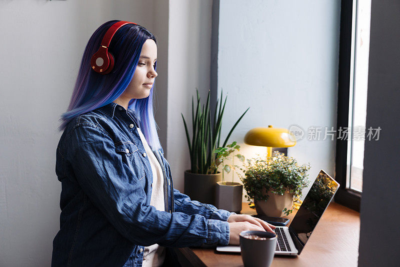 迷人的年轻拉丁裔女孩的肖像，蓝头发，蓝色牛仔衬衫，坐在咖啡馆里，在笔记本电脑上打字，还有复印空间。