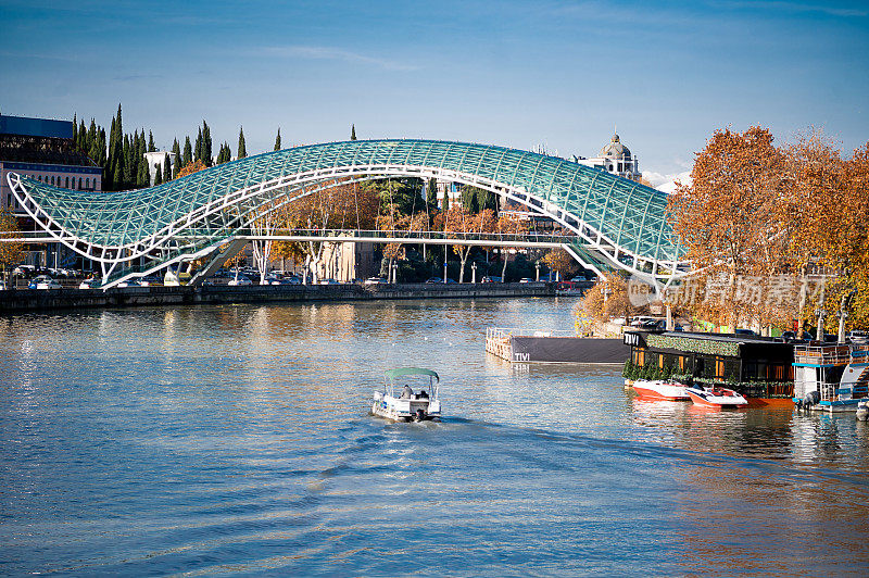 和平桥，一座现代建筑奇迹，横跨第比利斯的库拉河。河边的秋树增添了季节的魅力，船只在平静的水面上航行，