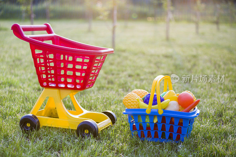 购物手推车和一个装着玩具水果和蔬菜的篮子。在阳光明媚的夏日，孩子们在户外玩的五颜六色的塑料玩具。