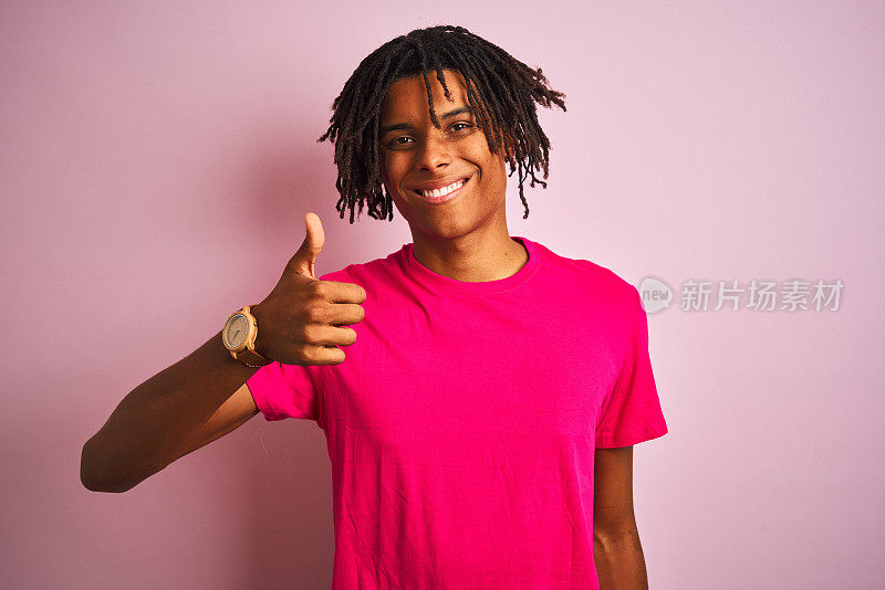 美国黑人男子与雷鬼发穿t恤站在孤立的粉红色背景做快乐的拇指手势与手。赞许的表情看着镜头，表示成功。