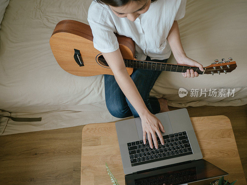 年轻女子微笑着坐在床上拿着吉他看着笔记本电脑