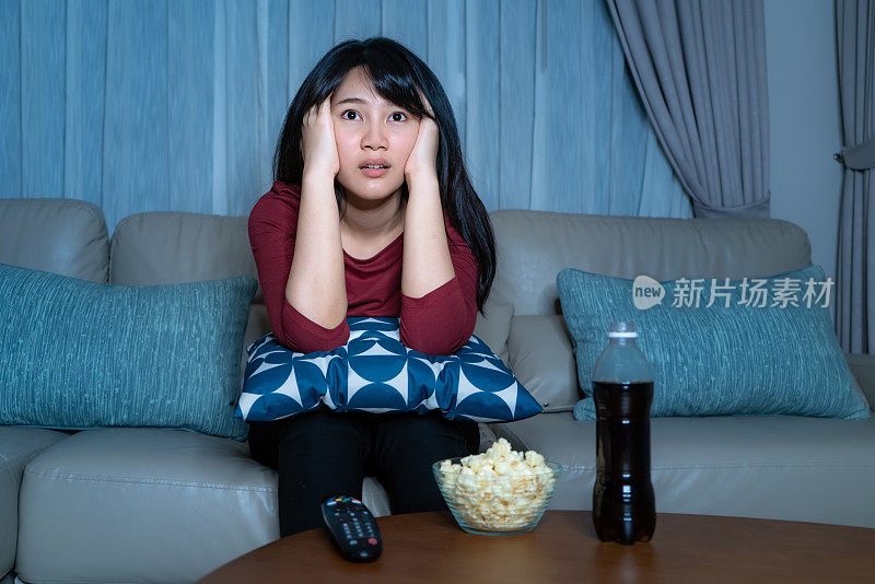年轻的亚洲女性看电视悬疑电影或新闻，看起来震惊和兴奋吃爆米花深夜在家里的客厅沙发上隔离。