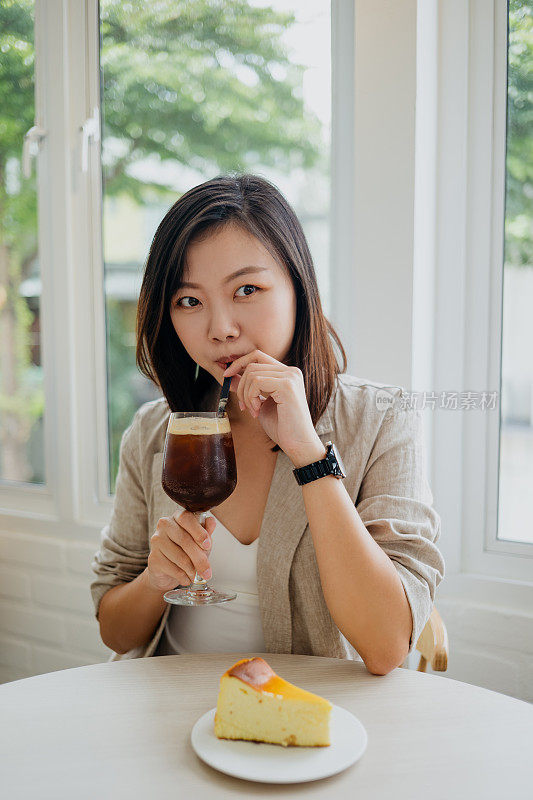 年轻的亚洲女人有一个冰咖啡饮料和芝士蛋糕在咖啡馆