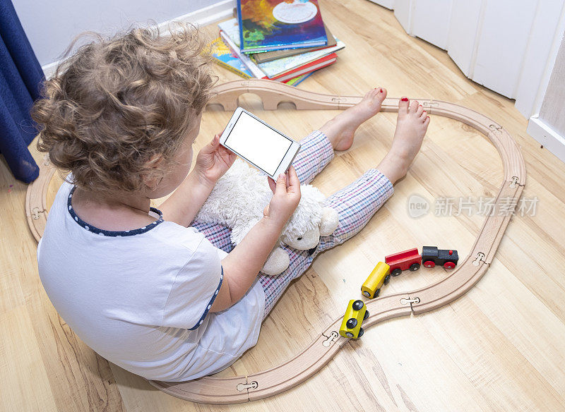 一个6岁的白人女孩坐在镶木地板上，手里拿着手机。可见的智能手机屏幕为复制空间