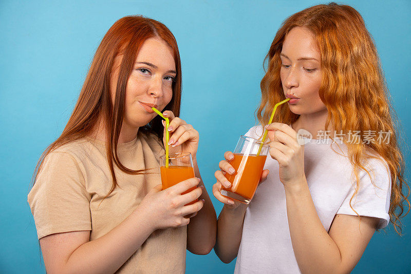 红发女友们穿着休闲，用吸管喝着橙汁。孤立在蓝色背景上。人们生活方式的概念。