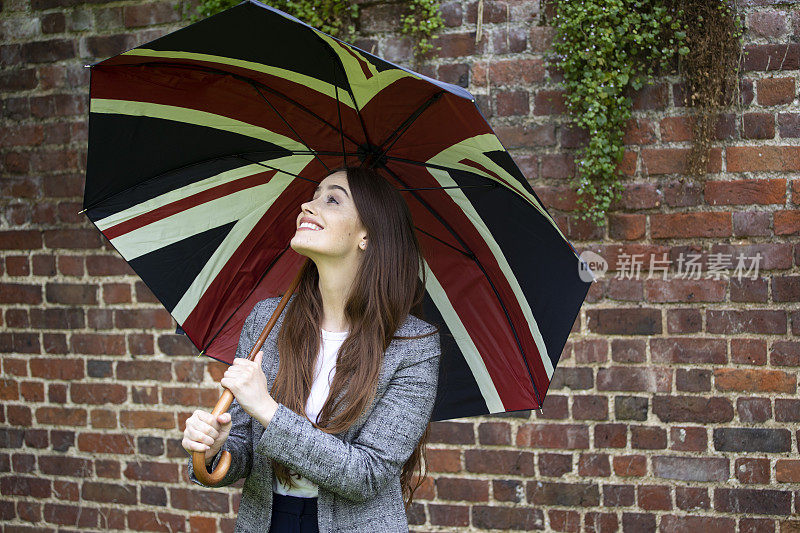 一位拿着英国国旗伞的年轻女子微笑着站在一堵砖墙旁。她穿着一件漂亮的灰色定制夹克。