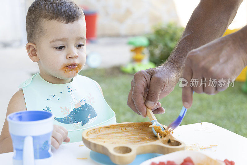 一个可爱的小男孩在他爷爷的帮助下吃饭。一个小男孩坐在花园里的椅子上吃东西。