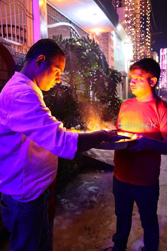 印度男人站在街上，一个男人拿着一个装有排灯节油灯的塔利(金属托盘)，另一个男人用火柴点燃灯芯，庆祝排灯节
