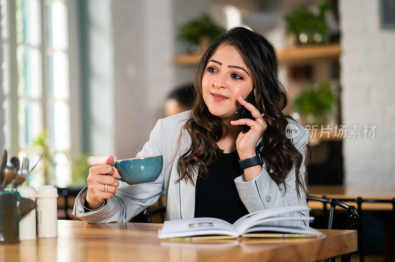 一名年轻女子一边使用智能手机，一边在咖啡馆看书、喝咖啡。