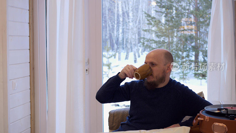 一个英俊的大胡子男人坐在家里的沙发上，用一个杯子喝着早晨的咖啡，他旁边是一个留声机，里面有一张黑胶唱片。这个人听音乐。