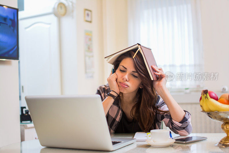 无聊的女学生用笔记本电脑做作业，头上顶着笔记本。