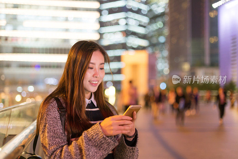 年轻美丽的亚洲女人晚上在现代城市玩手机