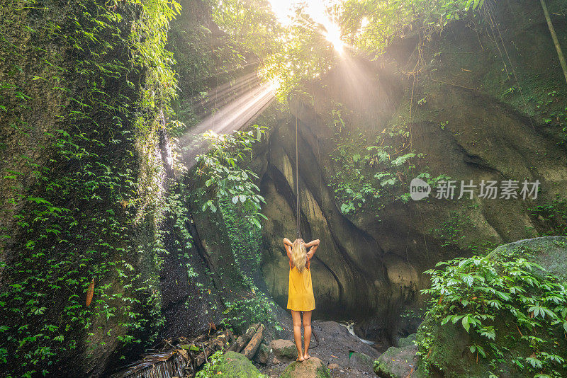 穿着黄色衣服的女孩站在热带雨林的洞穴里，仰望着从岩石上射下来的壮观的阳光。人们旅行的奇妙欲望自然的概念