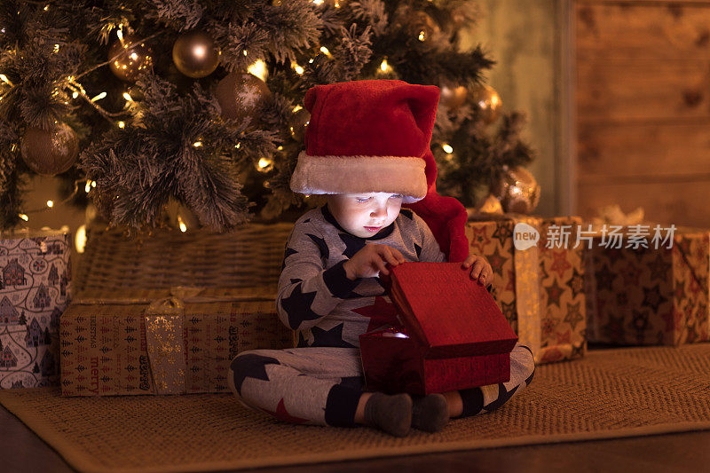 戴着圣诞老人帽子的快乐微笑的小男孩等待除夕打开礼物。圣诞节的概念与欢笑的孩子在圣诞树装饰花环和球