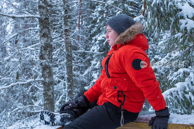 以白雪皑皑的森林为背景，一名身穿冬季红色夹克的白人青年望向远方。