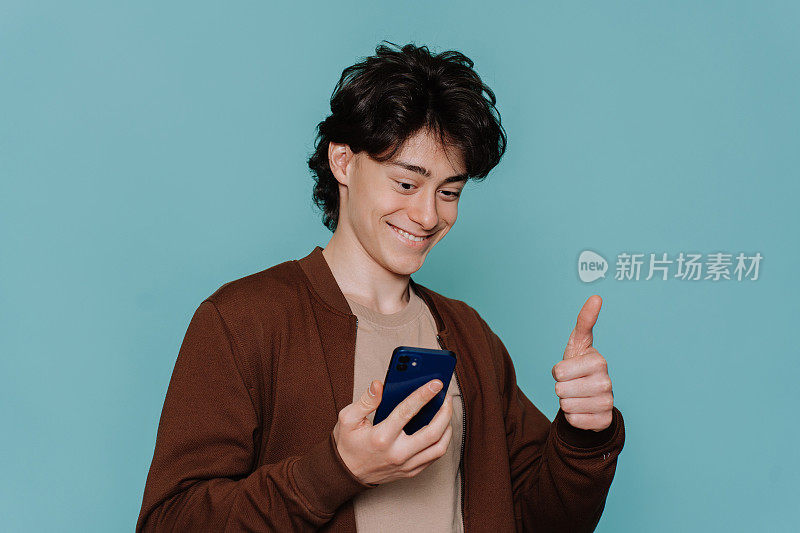 穿着棕色夹克的帅气男生在用智能手机进行视频通话时竖起大拇指，露出牙齿的微笑站在蓝绿色的演播室背景下。快乐的人得到好消息。