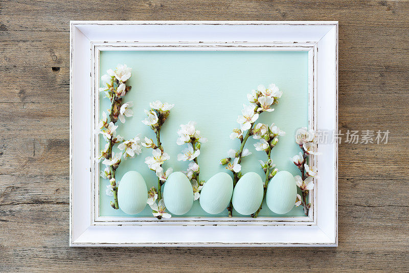 白色复古相框中摆放着薄荷绿色的复活节彩蛋，白色复古相框中的杏仁花位于乡村木制背景的中心