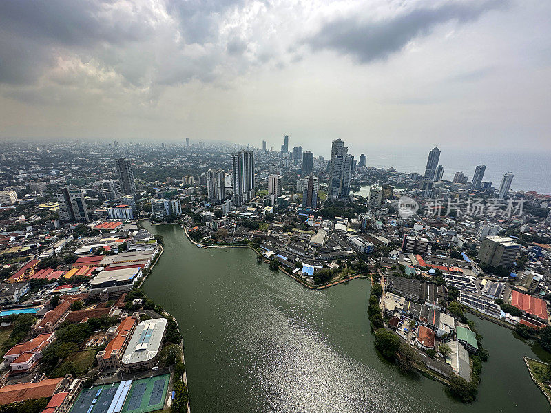 斯里兰卡科伦坡的城市景观图像，首都的摩天大楼，港口前，道路，铁路轨道，港口起重机，灰色多云的天空，重点在前景