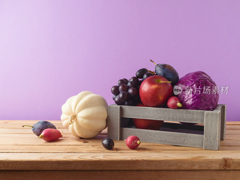秋收概念。南瓜、苹果、葡萄和蔬菜放在紫色背景的桌子上的木箱里