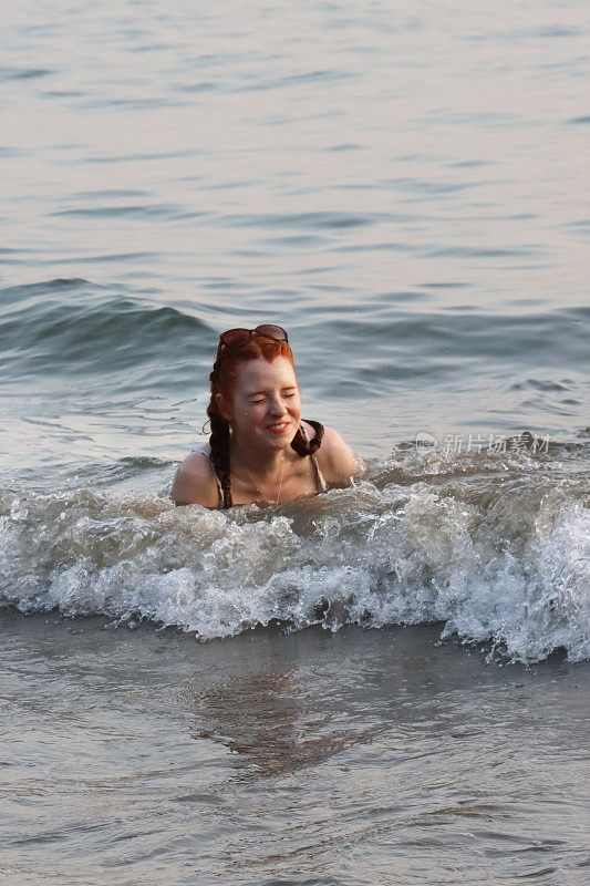 印度果阿，热带Palolem海滩，穿着两件式泳衣(比基尼)，一头红发、扎着辫子的女人在海滩度假时，掉进了浅滩，海浪拍打着她，拍打着海岸。