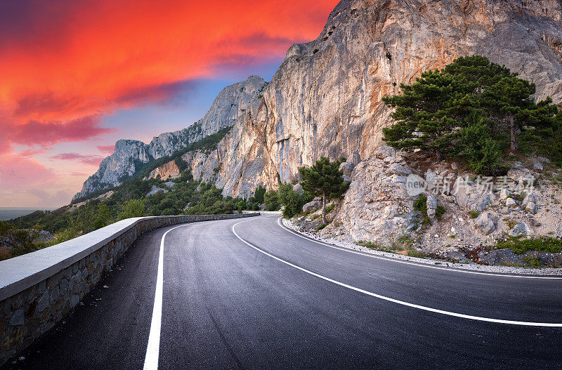 沥青道路。风景优美，蜿蜒的山路上有完美的柏油路面，高高的岩石，树木，惊人的天空和红色的云在夏季日落。全景。旅游的背景。山区高速公路