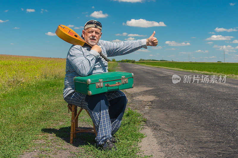 一位年长的搭便车者正带着一个古老的绿色手提箱和曼陀林在乡间路边休息