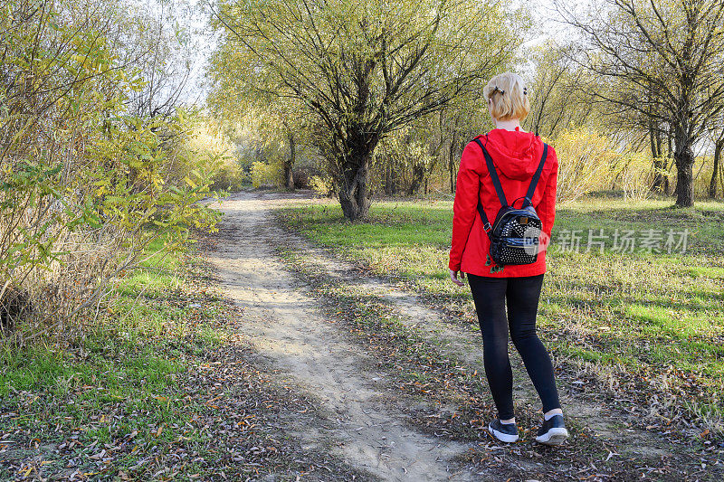 女孩穿着红色的夹克和一个背包沿着路去森林。路的开始。