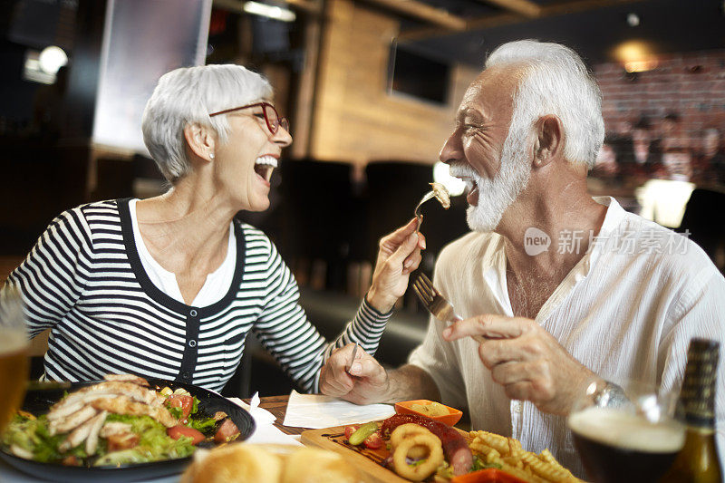 年长的女性笑着喂她的伴侣，而他们一起在餐厅吃饭