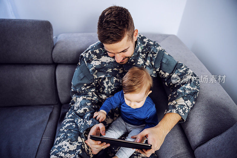 俯视图勇敢的士兵弥补失去的时间与他的儿子。一名男子抱着蹒跚学步的孩子，手里拿着平板电脑，看卡通片。