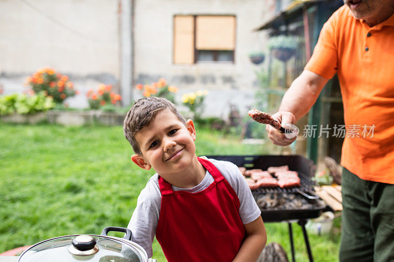 一个可爱的小男孩对着镜头微笑，他的爷爷正在他身后的烤架上给他烧烤