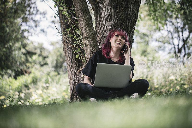 一个年轻的女人用笔记本电脑远程工作，她坐在公园的一棵树下。