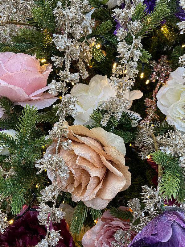 图像以白色、淡粉色、玫瑰金色和紫色玫瑰花冠装饰品挂在人造圣诞树的树枝上，云杉针叶上，模糊的白色神仙灯为背景
