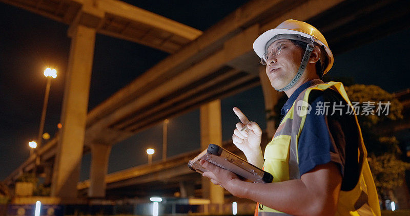 专家亚洲男土木工程师戴着安全帽、安全背心在工地上上夜班。工长在坚固的工业平板上检查结构进度和城市现场施工监督。