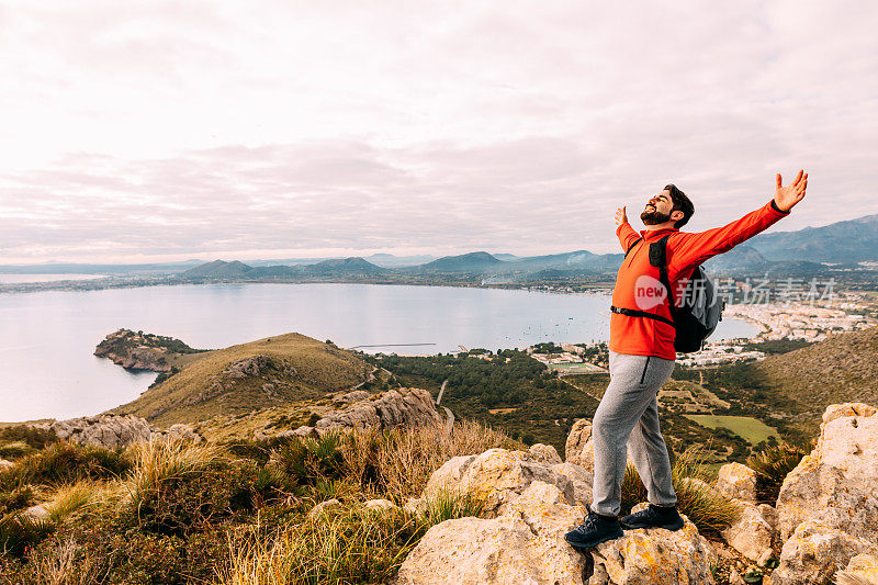 一名旅行者张开双臂站在马略卡岛北部海岸的风景前，背景是Pollensa湾