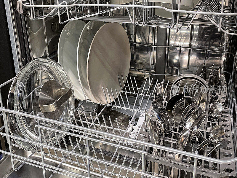 塑料洗碗机内部的特写图像，餐具篮里装着干净的不锈钢刀、叉和勺子，半满的机器里有干净的餐具，洗净晾干后就可以收起来，白色的餐盘，玻璃炖锅盖